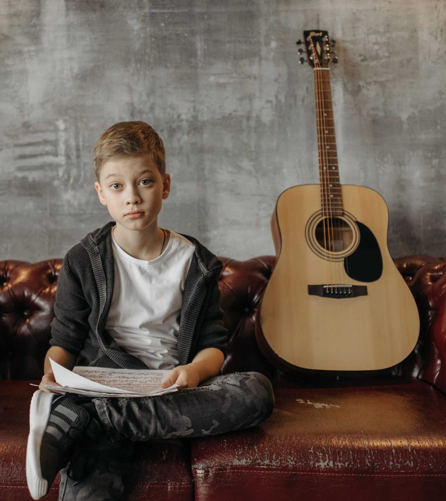 A young boy sitting on a leather sofa holding sheet music next to an acoustic guitar indoors.